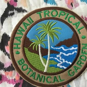 Puede incluir: Un parche bordado circular que muestra un paisaje tropical con dos palmeras, hierba verde, acantilados marrones y agua azul con olas blancas. El borde color canela contiene el texto bordado en verde HAWAII TROPICAL BOTANICAL GARDEN.