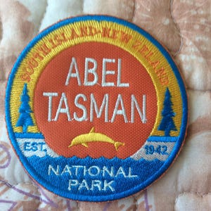 以下が含まれることがあります： 「ABEL TASMAN NATIONAL PARK」の文字とイルカのグラフィックが刺繍されたパッチ。パッチは、青い縁取りに「SOUTH ISLAND-NEW ZEALAND」と「EST. 1942」の文字が入っています。中央はオレンジ色です。