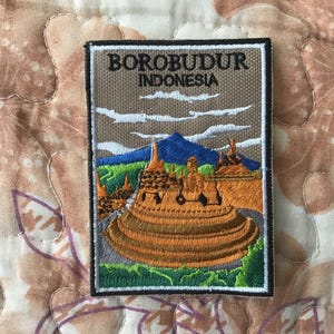 May include: Embroidered patch featuring the Borobudur temple in Indonesia. The patch has a rectangular shape with a black border. The design includes the text "BOROBUDUR INDONESIA", a mountain, clouds, and the temple in shades of orange, green, and blue.