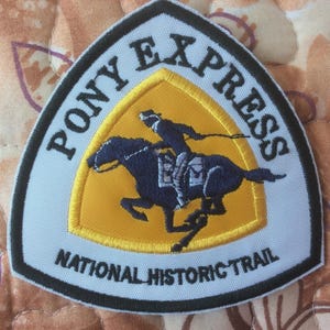 May include: A patch with the words "PONY EXPRESS" above a yellow triangle depicting a rider on a galloping horse. Below are the words "NATIONAL HISTORIC TRAIL". The patch has a white background and a black border.