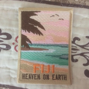 May include: Embroidered patch featuring a tropical beach scene with palm trees, ocean, and a sunset. The word "FIJI" is in orange, and "HEAVEN ON EARTH" is below. The patch has a beige border.