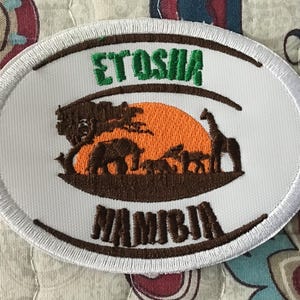 May include: An oval-shaped embroidered patch with a white background and a white stitched border. The design features a bright orange sun, brown silhouettes of an elephant, a giraffe, and other animals under a tree, with the text ETOSHA and NAMIBIA.