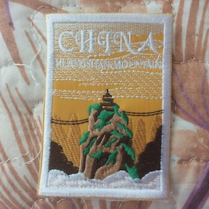 May include: Embroidered patch with a mountain scene, the text "CHINA" and "HUANGSHAN MOUNTAIN". The patch has a white border and depicts a mountain with green foliage, a pagoda, and a yellow sky.