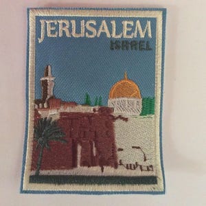May include: Embroidered patch depicting Jerusalem, with the text "JERUSALEM ISRAEL". The patch shows a cityscape with a golden dome, buildings, and a palm tree against a blue sky. It is bordered with light blue stitching.