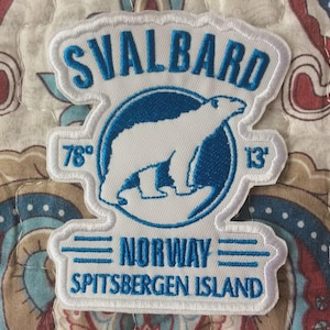 May include: Embroidered patch with a polar bear design, the text "SVALBARD" and "NORWAY SPITSBERGEN ISLAND". The patch includes the numbers "78°" and "13'" in blue. The patch has a white border. The patch is on a patterned background.