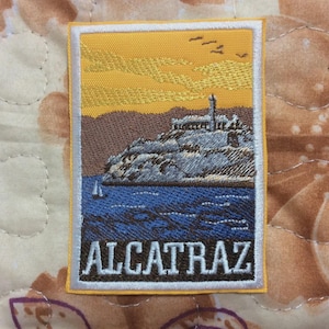May include: Embroidered patch featuring Alcatraz Island. The design includes a yellow sky, brown mountains, a gray island with a building, blue water, and the word "ALCATRAZ" in white letters.
