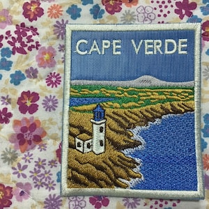 Puede incluir: Parche bordado con un paisaje costero con un faro, océano y montañas bajo un cielo azul. El texto "CAPE VERDE" está en la parte superior. El parche tiene un borde blanco cosido.