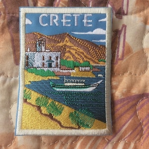 May include: Embroidered patch depicting a coastal scene with the word "CRETE" at the top. The design includes a white building, a boat, and a mountain range. The patch has a light blue border.
