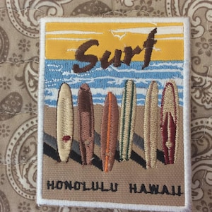 May include: Embroidered patch featuring a beach scene with the word "Surf" in brown script above a row of surfboards. The patch reads "Honolulu Hawaii" in black text below the surfboards. The background includes a yellow sky and blue ocean.