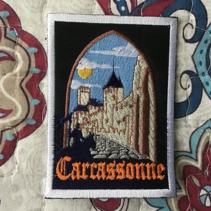 Op de afbeelding: Een rechthoekige geborduurde patch met een zwarte achtergrond en een witte rand. Het ontwerp toont een stenen kasteel in een gotische boog, een gele zon, een blauwe lucht en het silhouet van een ruiter. Het woord Carcassonne is in oranje.