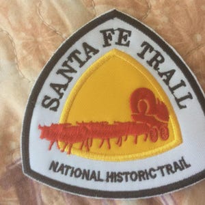 May include: Embroidered patch with the text "SANTA FE TRAIL" and "NATIONAL HISTORIC TRAIL". The patch features a yellow and red design of a covered wagon and cattle. The patch has a white background with a brown border.