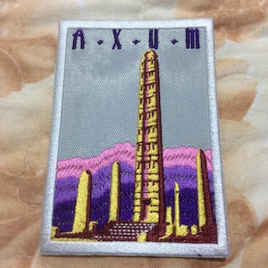 May include: Embroidered patch featuring a stylized depiction of a tall, ornate structure, possibly a monument or obelisk, in shades of yellow and brown against a gray background. The patch includes text at the top and is bordered by a white frame.