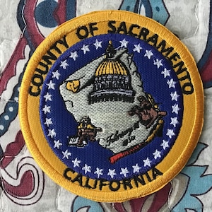 Op de afbeelding: Een ronde geborduurde patch met de tekst County of Sacramento California in zwarte letters op een gouden rand. Het midden is blauw met witte sterren en een witte kaart van de county met een koepelgebouw en kleine figuren.