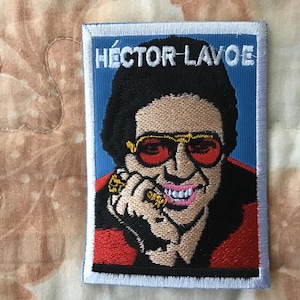 Könnte beinhalten: Ein rechteckiger bestickter Aufnäher mit einem Porträt von Héctor Lavoe vor blauem Hintergrund. Die Figur trägt eine rot getönte Sonnenbrille und eine rote Jacke, wobei der Name HÉCTOR LAVOE in weißen Buchstaben oben auf dem Aufnäher gestickt ist.