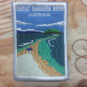 May include: Embroidered patch depicting the Great Barrier Reef in Australia. The design includes a blue sky with clouds, a green island, and a beach with a blue parasol. Text at the top reads "GREAT BARRIER REEF AUSTRALIA".