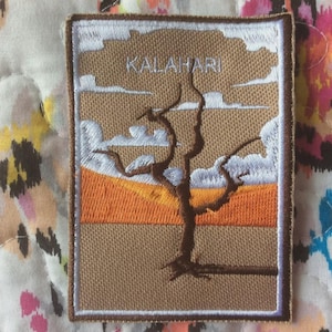 May include: Embroidered patch featuring a desert landscape with a bare tree and the word "KALAHARI" in white. The patch has a tan background, orange and yellow horizon, and white clouds. The patch is rectangular with a white border.