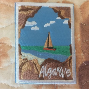 Puede incluir: Parche bordado que representa una escena costera con un velero en el agua bajo un cielo azul con nubes. El parche tiene un borde marrón y la palabra "Algarve" en escritura blanca.