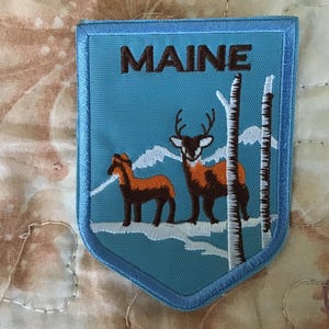 Pode incluir: Um emblema bordado em forma de escudo com fundo azul claro e a palavra MAINE em letras castanho-escuras. O design mostra dois veados castanhos num cenário nevado com montanhas brancas e duas bétulas brancas altas.
