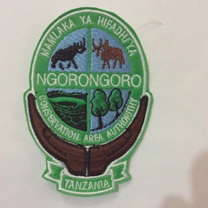 May include: Embroidered patch featuring the Ngorongoro Conservation Area Authority logo. The patch is green, brown, and blue, with text in English and Swahili. The design includes images of animals and landscapes, with the word "Tanzania" at the bottom.