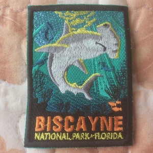 Puede incluir: Parche bordado con un tiburón martillo nadando en un océano turquesa. El parche tiene las palabras "BISCAYNE NATIONAL PARK FLORIDA" en letras naranjas. El parche es rectangular con un borde negro.