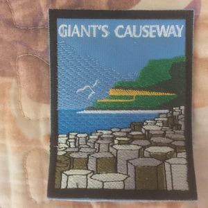 Puede incluir: Parche bordado con la Calzada de los Gigantes en Irlanda del Norte. El diseño incluye el texto "GIANT'S CAUSEWAY" sobre un paisaje costero con rocas hexagonales, agua azul y acantilados verdes. Dos aves blancas vuelan en el cielo.