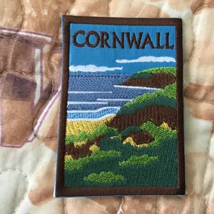 May include: Embroidered patch featuring the word "CORNWALL" above a coastal scene. The design includes a blue sky with white clouds, a blue ocean, green hills, and a yellow beach, all framed in a brown border.