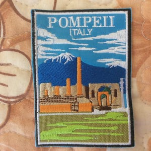 Puede incluir: Parche bordado con una escena de Pompeya, Italia. El parche tiene un fondo azul con nubes blancas y una montaña. El texto "POMPEII ITALY" está en la parte superior. Los edificios y estructuras están representados en naranja y beige, con hierba verde en la parte inferior.