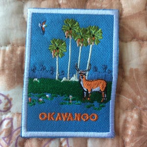 Może przedstawiać: Haftowana naszywka przedstawiająca krajobraz z palmami, jeleniem i ptakiem na tle błękitnego nieba. Słowo "OKAVANGO" jest wyhaftowane na pomarańczowo. Naszywka ma białą obwódkę.