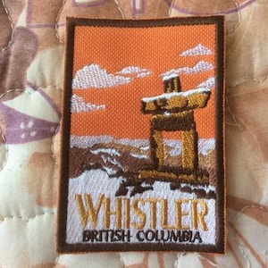 Puede incluir: Parche bordado con un tótem marrón y dorado en un paisaje nevado bajo un cielo naranja con nubes blancas. El parche dice "WHISTLER BRITISH COLUMBIA" en letras doradas y blancas, enmarcado por un borde marrón.
