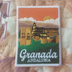 以下が含まれることがあります： 夕焼けのグラナダ、アンダルシアの風景が描かれた刺繍パッチ。パッチは長方形で白い縁取りがあります。「Granada ANDALUSIA」の文字が緑色の背景に白で刺繍されています。