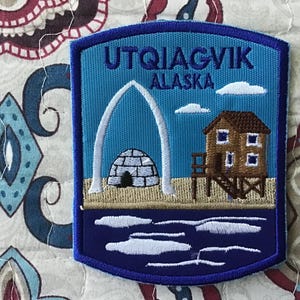 Puede incluir: Un parche bordado rectangular con borde azul que presenta el texto UTQIAGVIK ALASKA. El diseño muestra un arco de hueso de ballena blanco, un iglú gris y una casa de madera sobre pilotes sobre un suelo color canela con agua azul oscuro.