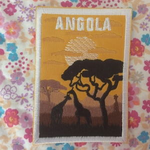 May include: Embroidered patch featuring an African savanna scene with giraffes, trees, and a sunset. The word "ANGOLA" is stitched in white across the top. The patch has a white border and is set against a floral background.
