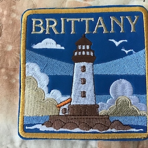 May include: Embroidered patch featuring a lighthouse with the word "BRITTANY" above it. The patch has a blue background with white clouds, seagulls, and a yellow border. The lighthouse is white and brown.