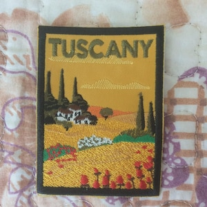 May include: Embroidered patch featuring a Tuscan landscape with the word "TUSCANY" at the top. The design includes rolling hills, cypress trees, and a field of red flowers, all set against a yellow background.