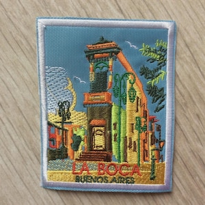 May include: Embroidered patch depicting La Boca, Buenos Aires. The design shows buildings, a tower, and the text "LA BOCA BUENOS AIRES" in gold lettering, set against a blue background with a white border.