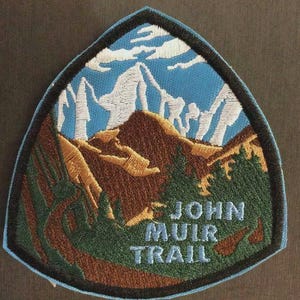 Könnte beinhalten: Gestickter Aufnäher mit einer Berglandschaft und dem Text "JOHN MUIR TRAIL". Der Aufnäher hat einen blauen Rand und eine dreieckige Form. Das Design umfasst Berge, Bäume und Wolken in Braun, Grün, Weiß und Blau.