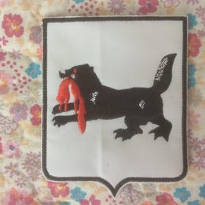 Może przedstawiać: Haftowana naszywka przedstawiająca czarne zwierzę trzymające czerwony przedmiot w pysku, na białym tle w kształcie tarczy. Naszywka ma ozdobną obwódkę. Tło to wzór kwiatowy.