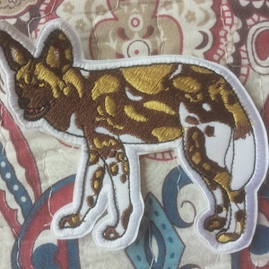 May include: Embroidered patch featuring a stylized African wild dog. The patch has a white border and depicts the dog in shades of brown, gold, and white. The patch is likely for clothing or accessories.