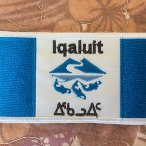 Peut inclure: Un écusson rectangulaire avec un centre blanc et des bandes bleues verticales de chaque côté. Le centre présente le mot "Iqaluit" en noir, au-dessus d'un dessin bleu de montagne et de rivière, et du texte dans une autre écriture en dessous.