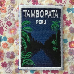 May include: Embroidered patch featuring the text "TAMBOPATA PERU" in white. The patch depicts a dark blue night scene with palm trees and a silhouette of a jungle landscape. The patch has a white border.