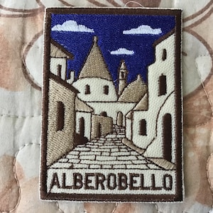 Puede incluir: Parche bordado con una ilustración detallada de Alberobello, Italia. El diseño muestra una calle adoquinada bordeada de casas trullo tradicionales, bajo un cielo azul con nubes blancas. La palabra "ALBEROBELLO" se muestra en la parte inferior.