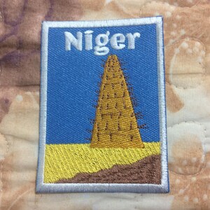 May include: Embroidered patch with the word "Niger" in white above a tan structure on a blue background. The structure is on a yellow and brown landscape, framed by a white border. A travel souvenir.