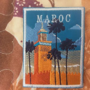 Può includere: Toppa ricamata raffigurante una scena marocchina. La toppa ha uno sfondo blu con la scritta "MAROC" in bianco. Raffigura un alto edificio arancione e giallo con un minareto, palme e una scena di spiaggia.