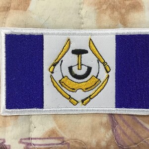May include: A rectangular embroidered patch featuring a vertical triband design of blue, white, and blue. The central white section displays a gold-coloured emblem consisting of four crossed tools surrounding a central black and gold circular icon.
