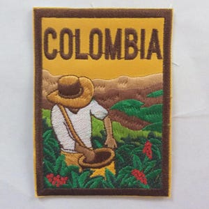 Op de afbeelding: Geborduurde patch met het woord "COLOMBIA" boven een illustratie van een koffieboer. De patch heeft een gele achtergrond met bruine en groene details en een bruine rand. Het ontwerp toont een koffieplantage.