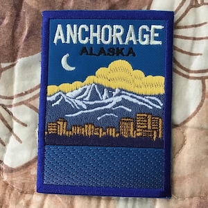 May include: Embroidered patch featuring the text "ANCHORAGE ALASKA" in white. The patch depicts a landscape with mountains, a crescent moon, and a cityscape. The patch has a blue border and background.