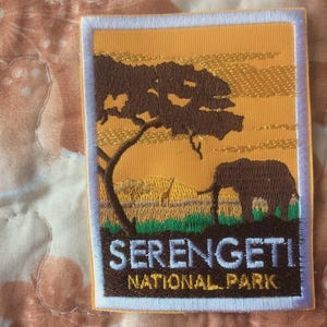 May include: Embroidered patch with a Serengeti National Park design. It shows a brown tree, an elephant, and a giraffe against a yellow and orange sunset. The words "SERENGETI NATIONAL PARK" are embroidered in brown and white.