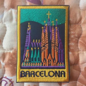 Może przedstawiać: Haftowana naszywka przedstawiająca Sagrada Familia w Barcelonie. Naszywka ma czarne tło z katedrą w kolorze pomarańczowym, fioletowym i zielonym. Słowo "BARCELONA" jest w kolorze żółtym na dole, z żółtą obwódką.