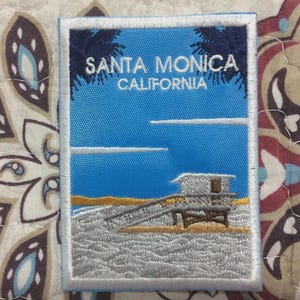 Puede incluir: Parche bordado con una escena de playa con un puesto de salvavidas, olas del océano y el texto "SANTA MONICA CALIFORNIA" sobre un cielo azul. El parche tiene un borde blanco, ideal para ropa o bolsos.