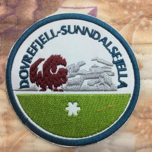 May include: Circular embroidered patch with a blue border and the text "DOVREFJELL-SUNNDALSEJELA". The patch depicts a maroon animal, grey logs, and a green field with a white asterisk and a gold line.